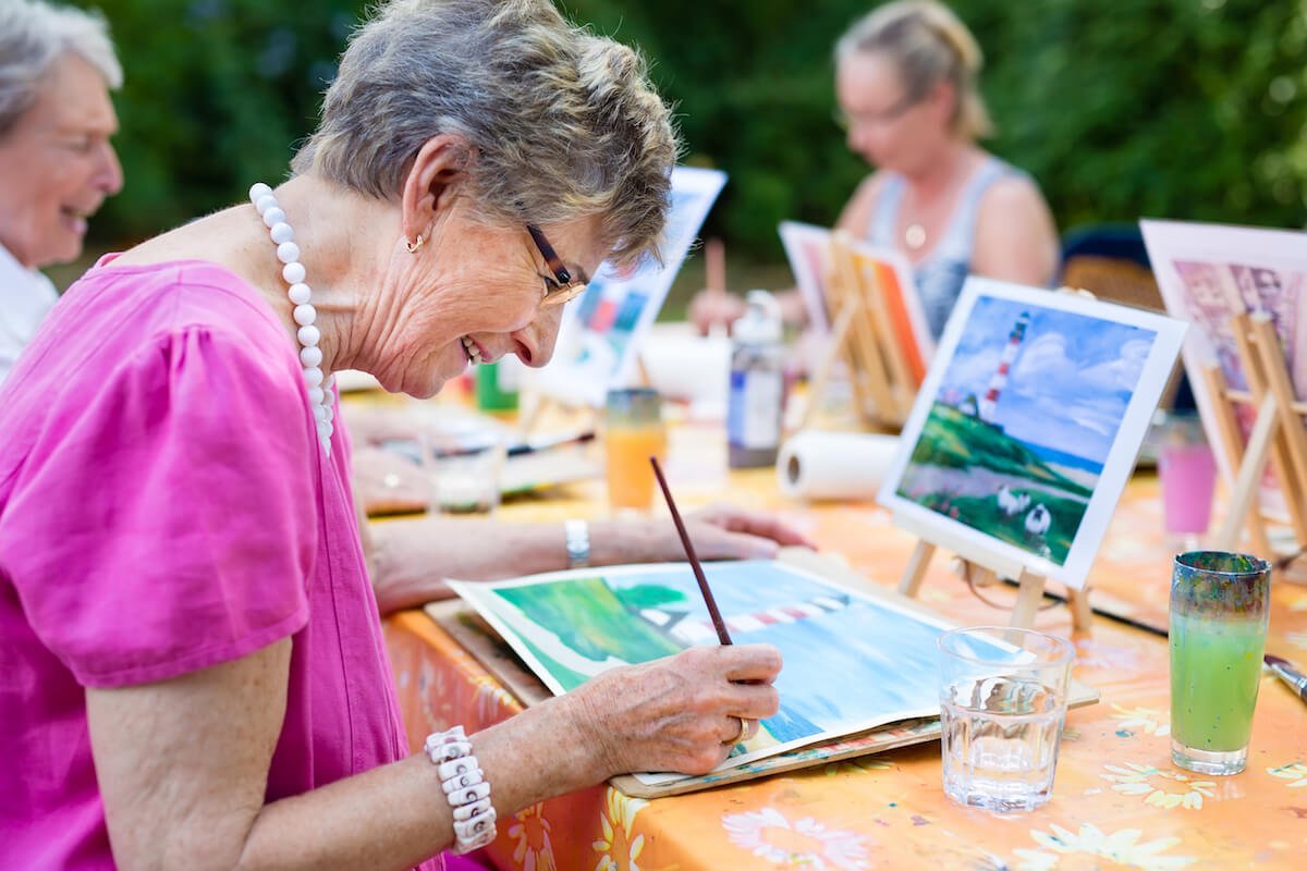 Side view of a happy senior woman smiling while drawing as a recreational activity or therapy outdoors together with the group of retired women.