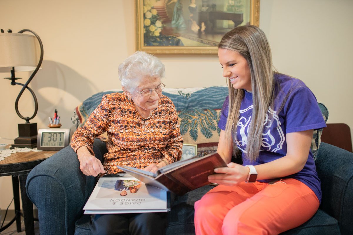 Smiling Senior Woman and Caregiver Look at Books While Sitting on Couch_The Pavilion Senior Living